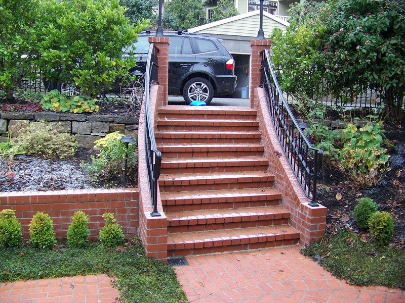 Brick outdoor staircase with black metal railings leading to a higher level with a parked black car. The surrounding area features landscaped greenery, including small shrubs and a stone retaining wall.