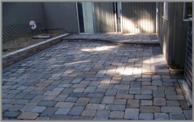 A brick pavere patio with rectangular and square gray stones arranged in a pattern. The patio leads to a sliding glass door on a gray building. There's a small raised area with a retaining wall on the left side.