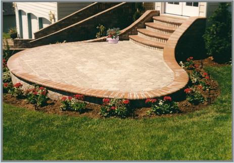 A curved stone patio with brick edging is bordered by a well-maintained flower bed featuring red and pink flowers. The patio is connected to a set of wide brick stairs leading up to a house, with lush green grass surrounding the area.