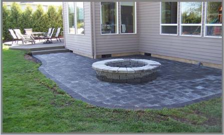 A backyard patio featuring a circular stone fire pit at the center, surrounded by gray pavers. The patio is adjacent to a house with beige siding and multiple windows. In the background, there is a table with chairs and a lawn area bordered by greenery.