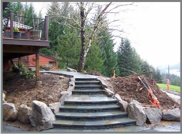 A stone staircase leads up a small hill surrounded by large rocks and soil. A wooden deck with a railing is visible on the left. In the background, there are tall evergreen trees and a glimpse of a distant landscape. The sky is overcast.