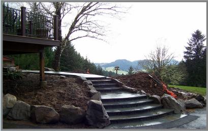 A stone staircase leads up a small slope, bordered by large rocks. To the left, there's part of a wooden deck, and in the background, there are trees and a view of distant hills under an overcast sky.
