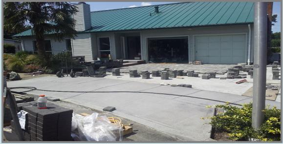 A house with a green metal roof is having a driveway renovation. The driveway is partially paved with concrete and stone pavers. Construction materials, including stacked bricks and bags, are visible in the foreground. The garage is open, suggesting ongoing work.