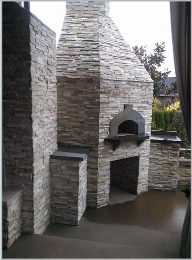 Outdoor stone fireplace with a tall chimney and arched baking oven, set on a patio.