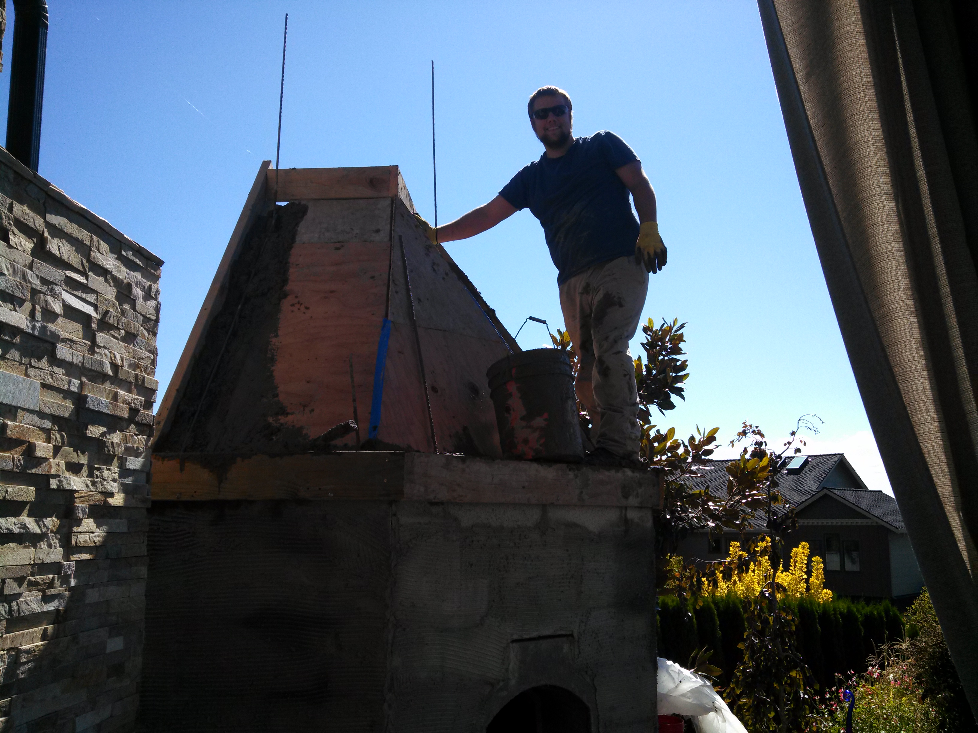 A man standing on a wooden platform working with a large stone structure, with a clear blue sky and trees in the background.