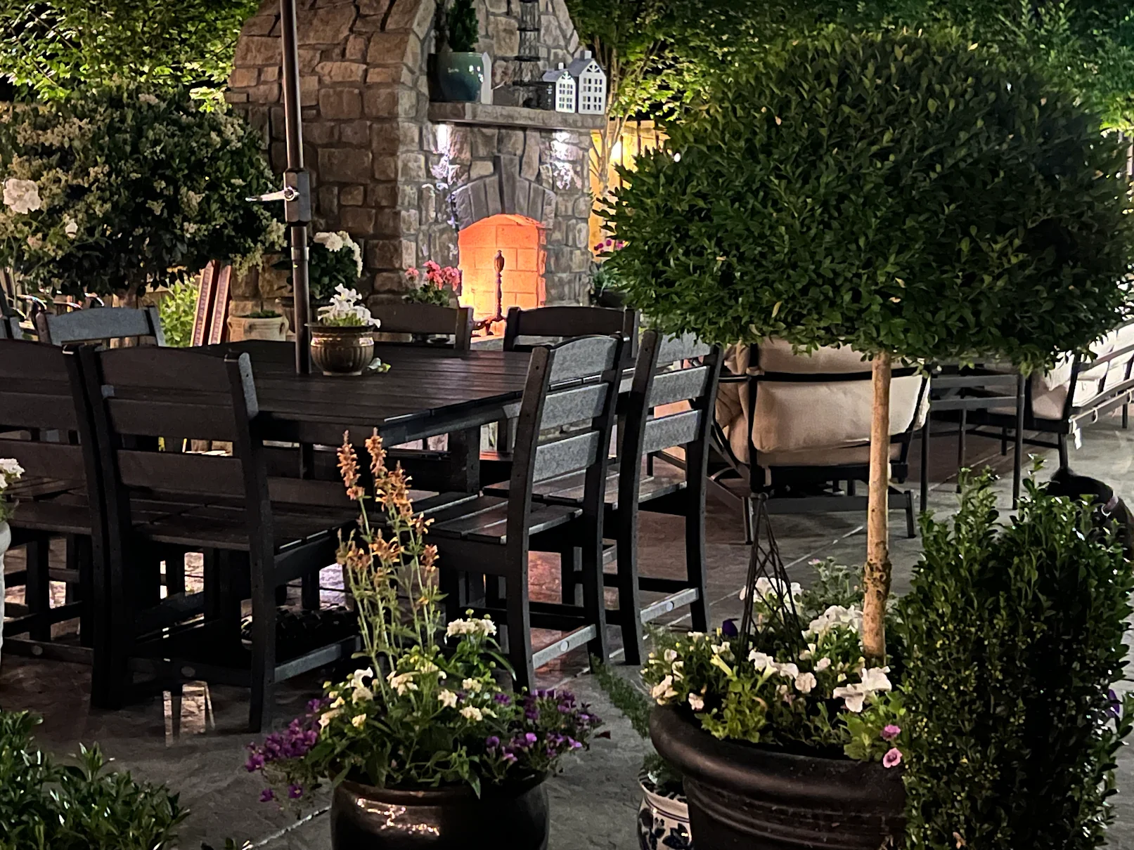 A cozy outdoor patio at night with a lit stone fireplace. Soft seating and an umbrella are on the left, surrounded by illuminated string lights and potted plants.